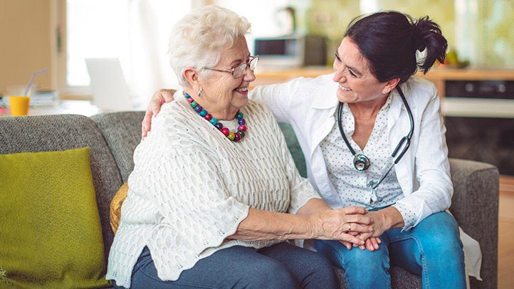 a home health care worker and an elderly client smile at each other while sitting on a sofa