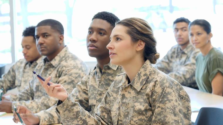 six service members in uniform sit at tables in a classroom