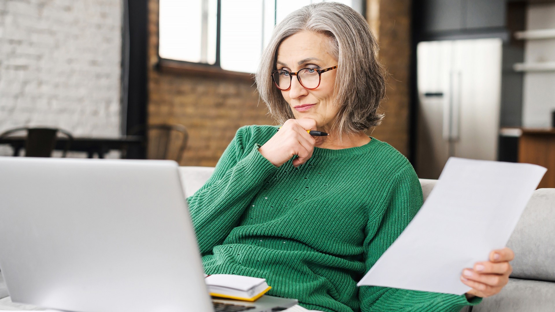a woman with gray hair and glasses holds papers as she thinks about the information she's seeing on a laptop screen