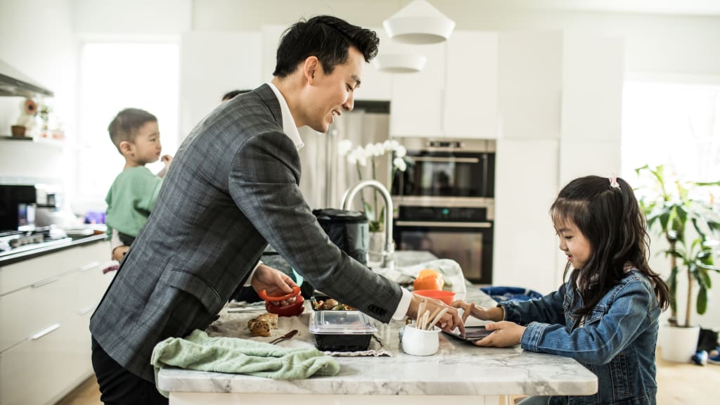 a busy father wearing a suit entertains his school-age daughter at the kitchen island as a mother holds a young child in the background