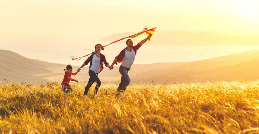 a smiling family of mother, father, and small child runs through a field as the father holds a kite aloft
