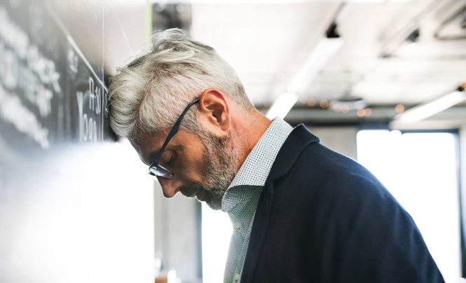 a white-haired professional leans his head against an office wall in discouragement