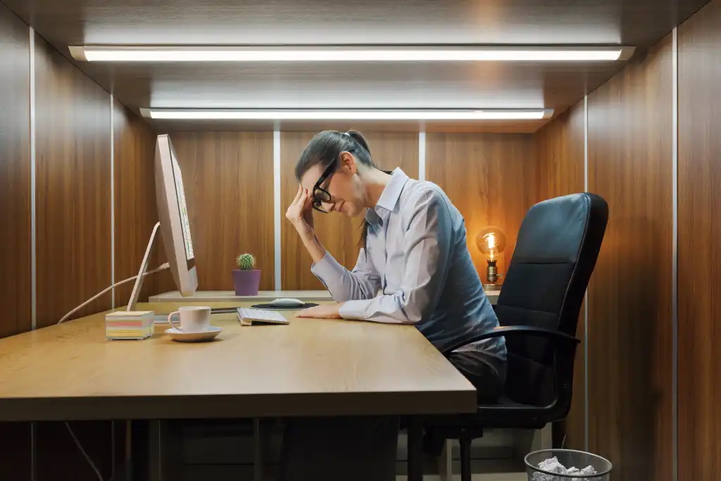 a frustrated worker sits in a tiny cubicle with fluorescent lights overhead