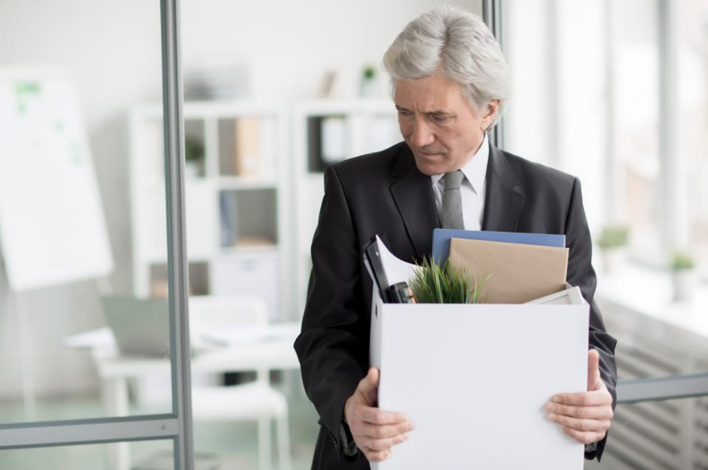 a gray-haired executive looks back sadly as he leaves his office with a box of belongings
