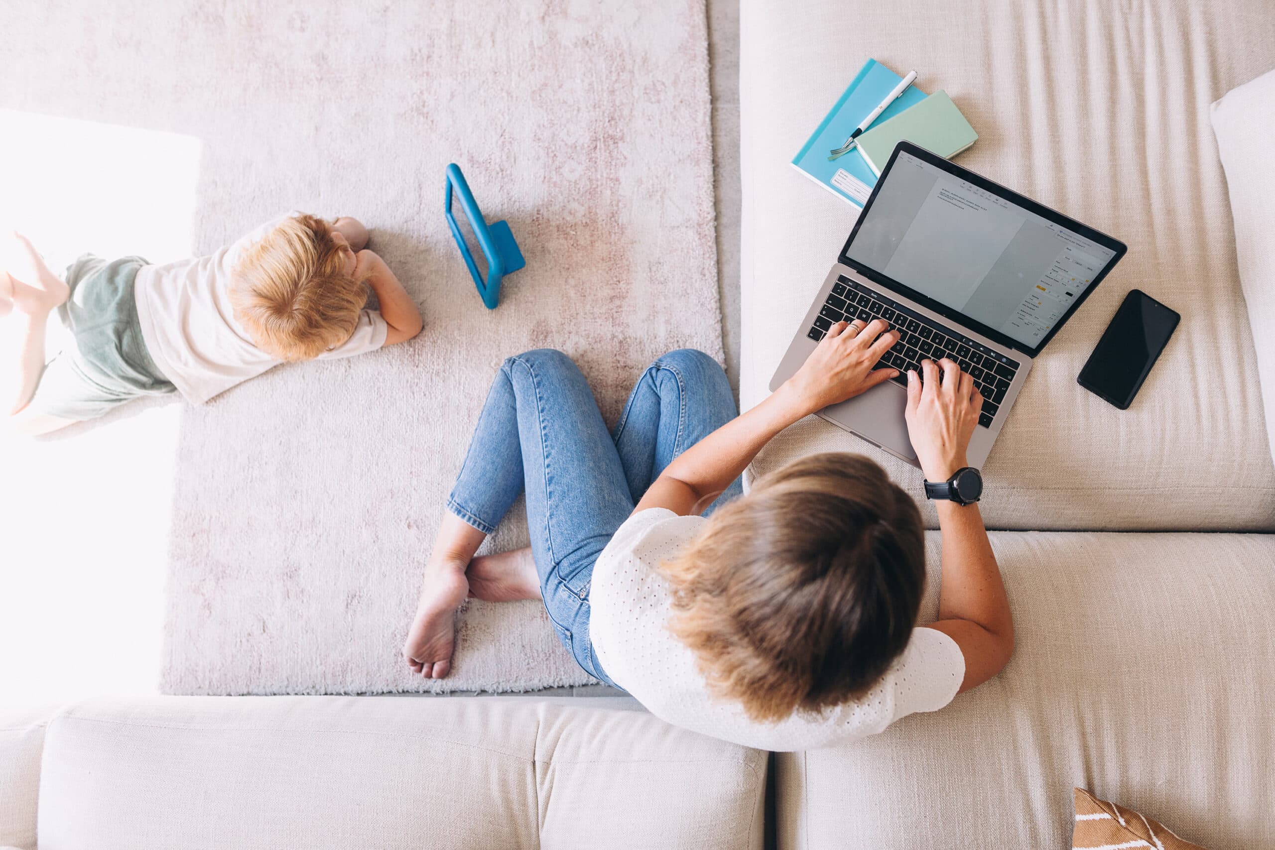 a mom sits on her living room floor, working on a business on her laptop, while her child watches a video on a tablet computer next to her