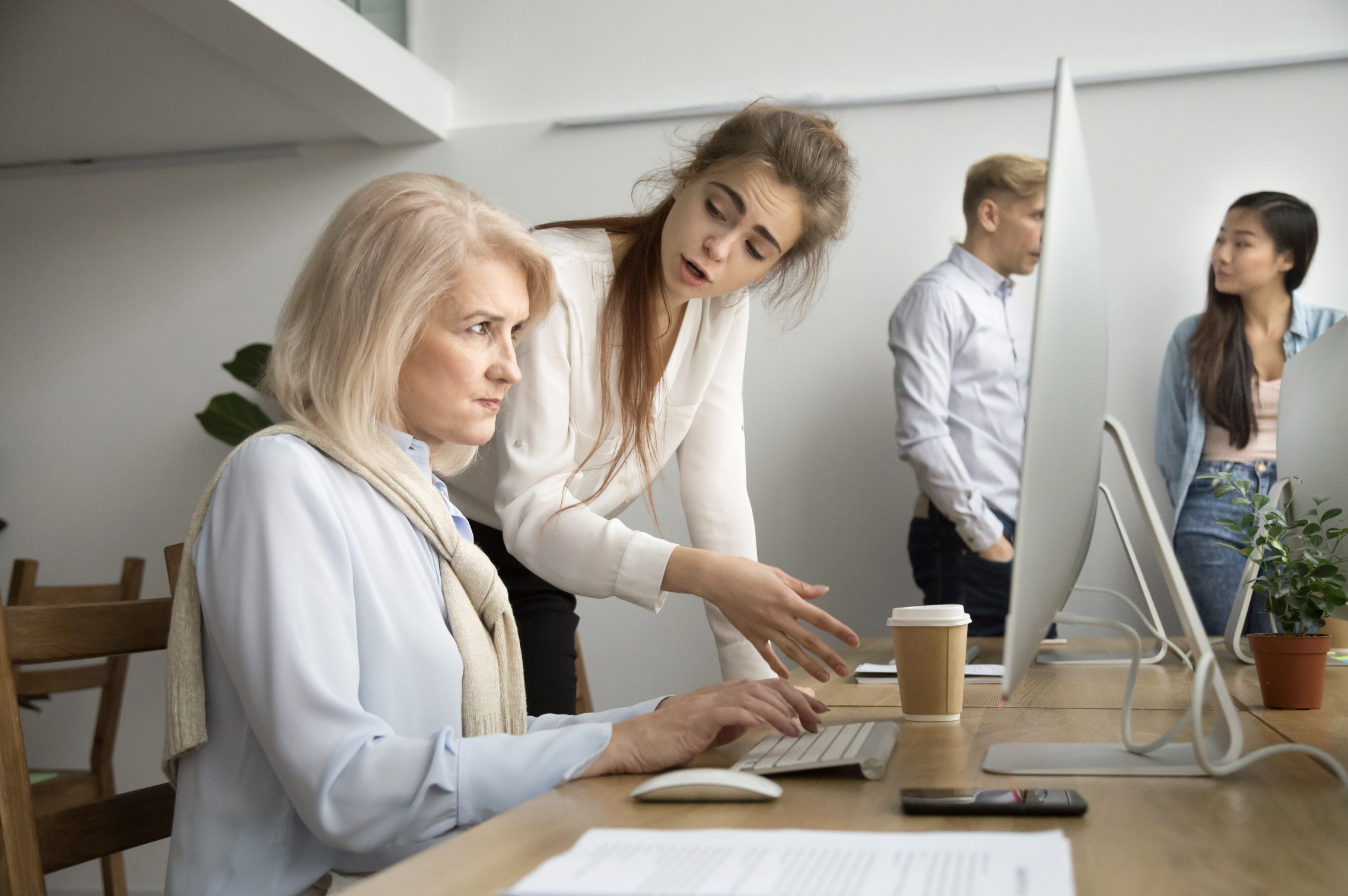a younger worker condescendingly points out something on a computer screen to a frustrated older worker