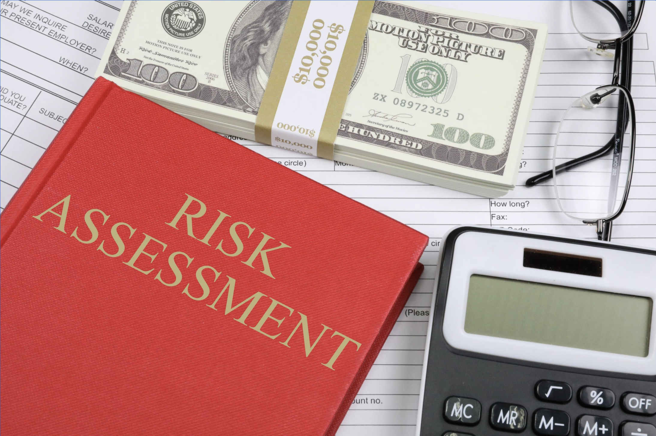 a red book titled "risk assessment" sits on top of financial forms next to a stack of 100-dollar bills, a calculator, and reading glasses